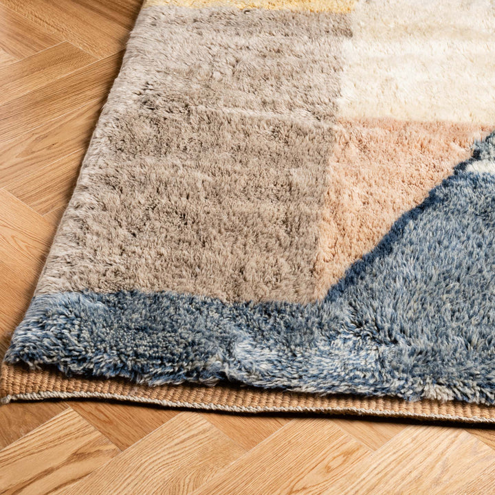 Close-up of a handmade Moroccan wool rug with abstract design and shag texture, featuring earthy and blue tones on wooden flooring.
