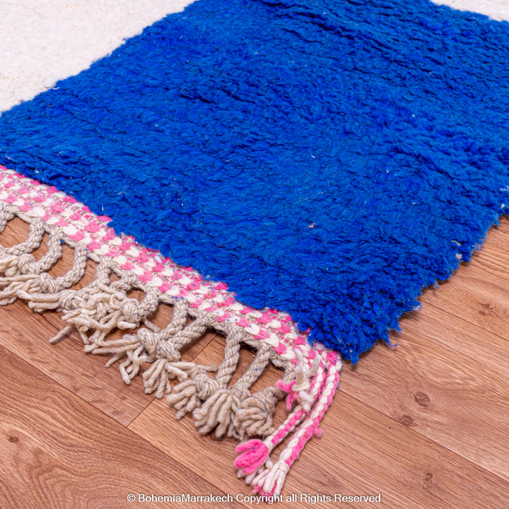 Handmade blue Moroccan wool rug with pink fringe on wooden floor.