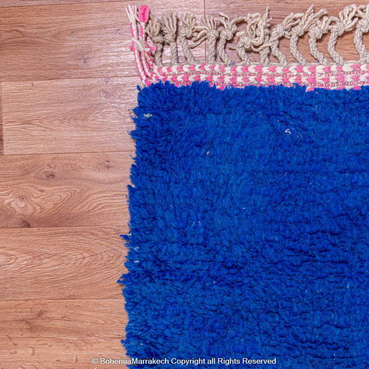 Corner view of a blue handmade Moroccan wool shag rug with tassel fringe on hardwood floor.