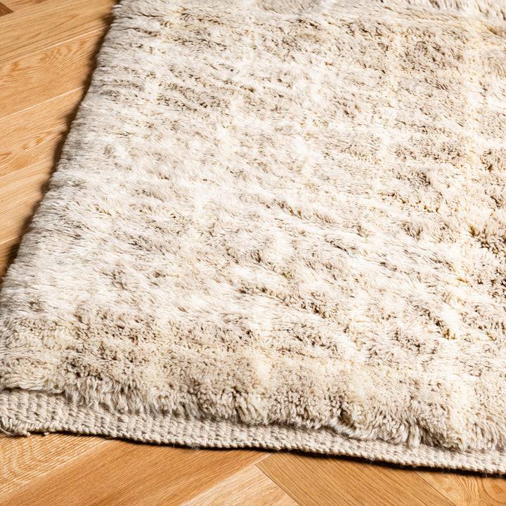 Beige Moroccan wool rug with subtle grid pattern on wooden floor, handmade and soft, ideal for minimalist interiors.