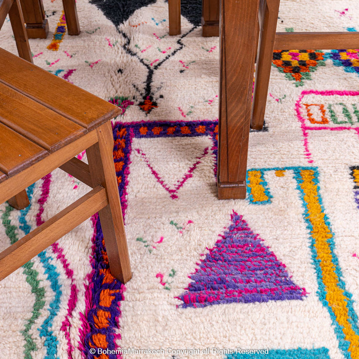 Colorful Azilal Moroccan Berber rug with tribal motifs under a wooden table, showcasing vibrant pink, blue, and orange patterns.
