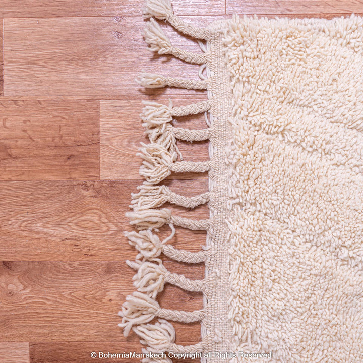 Cream Moroccan Berber rug on wooden floor, showcasing tassel detail. Handmade wool area rug from Morocco.