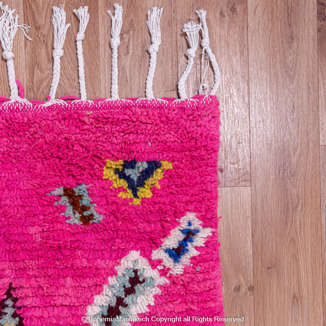 Vibrant pink Moroccan Berber rug with colorful motifs and fringe on a wooden floor.
