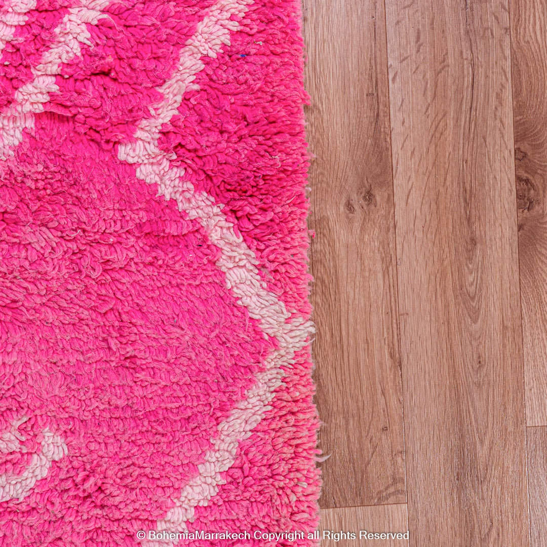 Vibrant pink Moroccan rug with white diamond motifs on wood floor, handwoven by Berber artisans using natural wool.