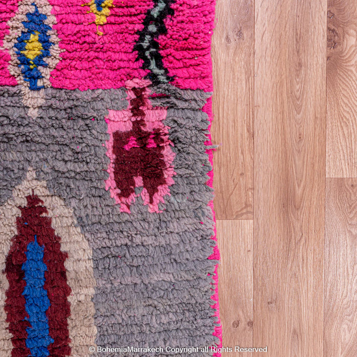 Close-up of a vibrant pink Moroccan Berber rug with traditional motifs on a wooden floor, featuring colors like gray, blue, and red.
