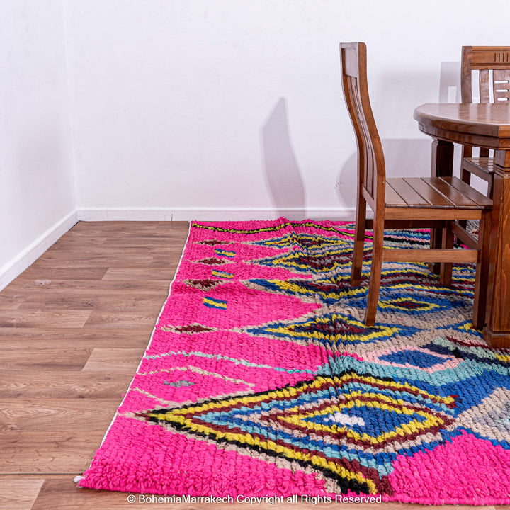 Pink Moroccan rug with vibrant multicolor Berber patterns under a wooden dining table, handmade with natural wool.