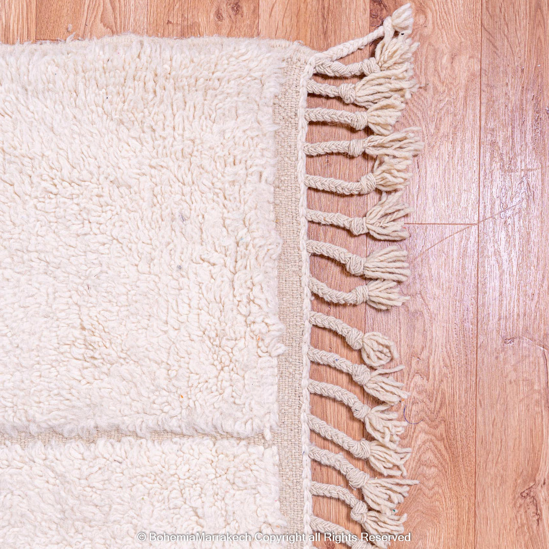 White Moroccan wool rug with geometric grid and fringes on wood floor, modern Beni Ourain style, handmade by Berber artisans.