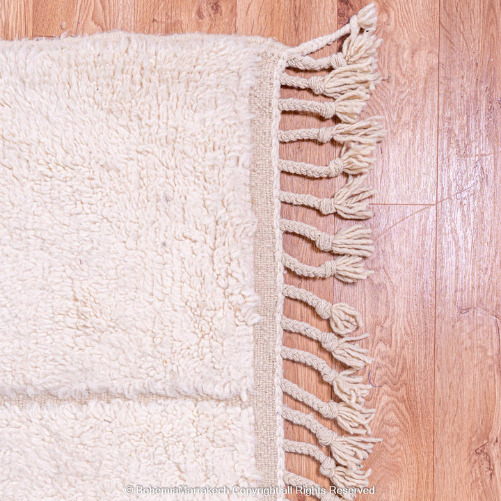 White Moroccan wool rug with geometric grid and fringes on wood floor, modern Beni Ourain style, handmade by Berber artisans.