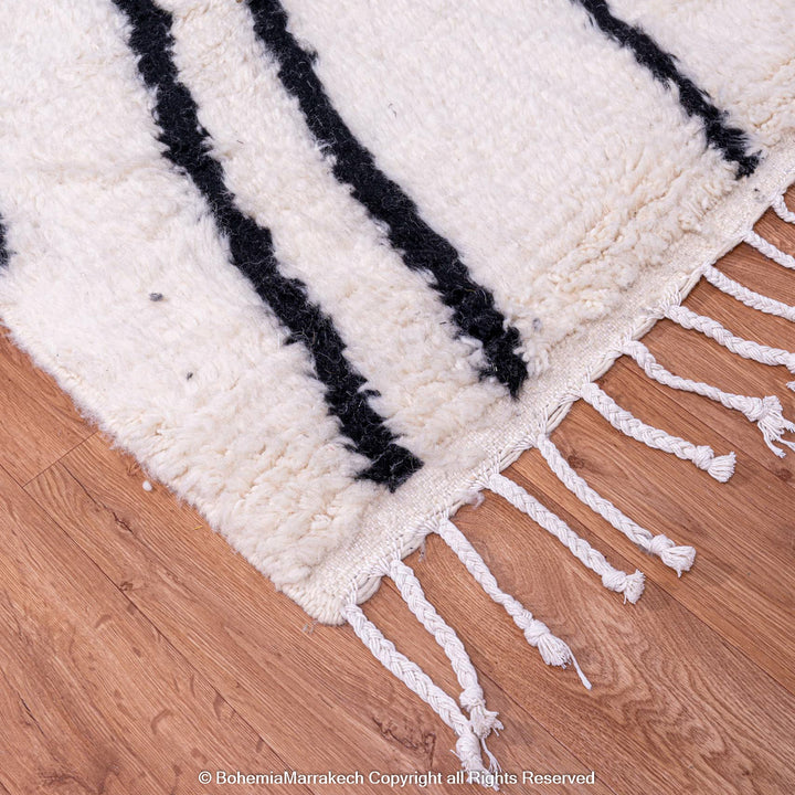 Black and white Beni Ourain Moroccan wool rug with black criss-cross patterns on a wooden floor, showing fringe detail.