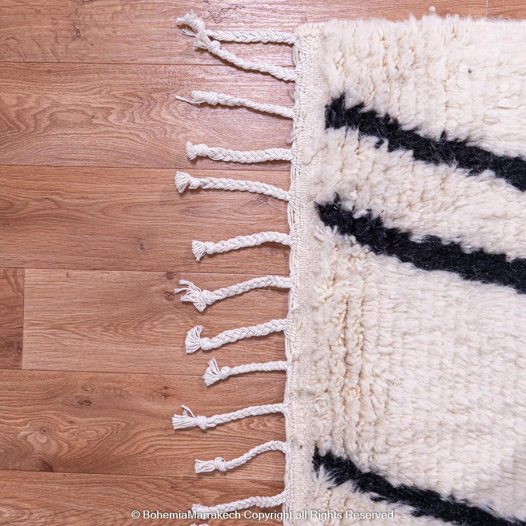 Close-up of Beni Ourain Moroccan wool rug with black abstract patterns, showcasing tassels on natural wood floor.