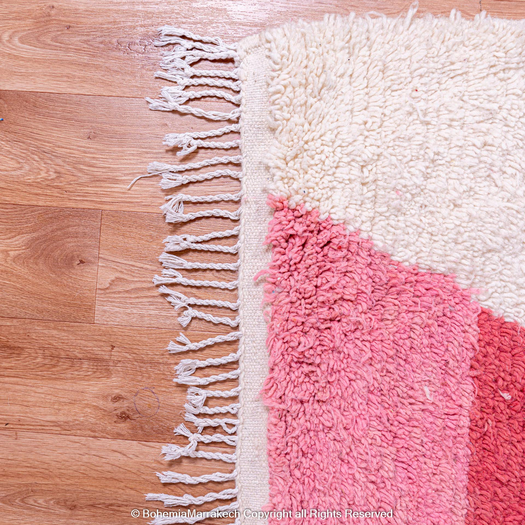 Handmade Moroccan wool rug in pink and red, featuring hourglass design, natural base, and fringe detail. Perfect for modern interiors.