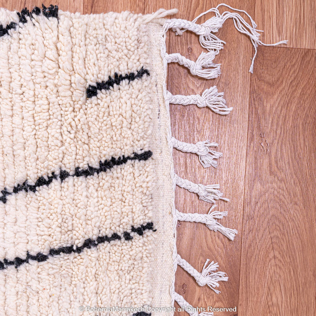 Close-up of a white Moroccan wool rug with black abstract diamond patterns and fringes on a wooden floor.