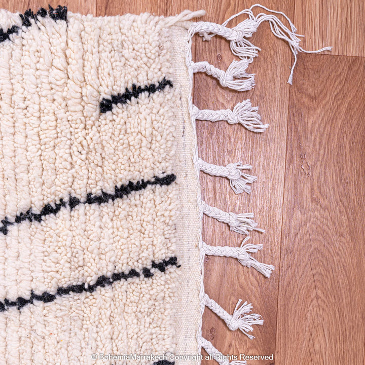 Close-up of a white Moroccan wool rug with black abstract diamond patterns and fringes on a wooden floor.