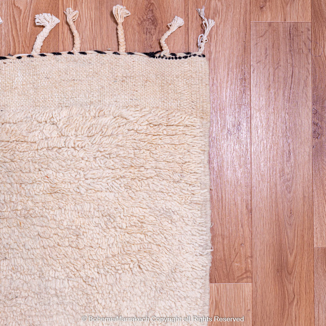 Details of a cream Moroccan wool rug with fringed edges on a wood floor, showcasing texture and craftsmanship.