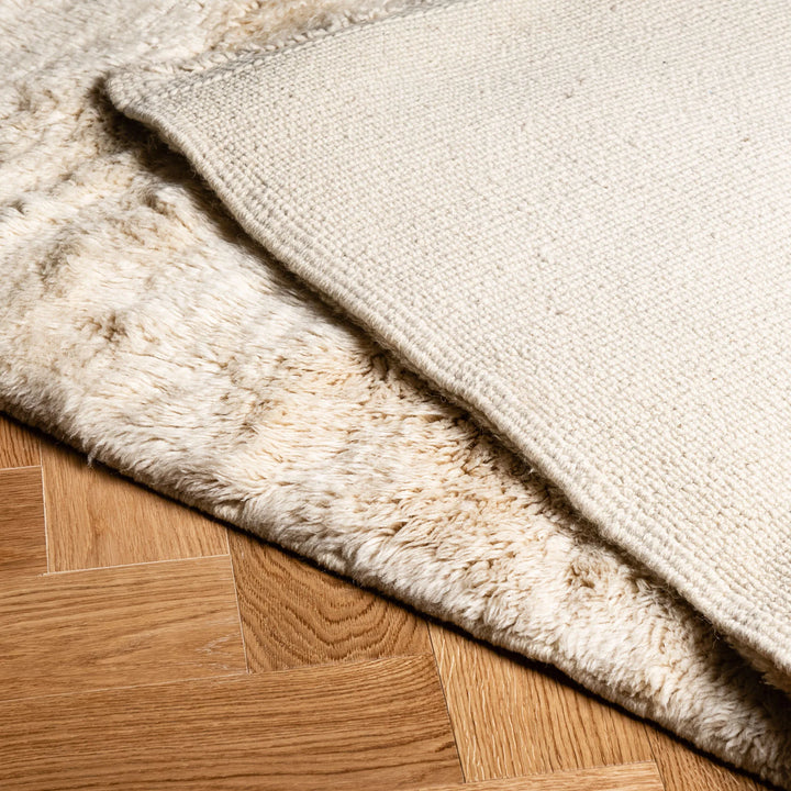 Close-up of a plush, handmade Moroccan rug in beige and ivory tones on a wooden floor, showcasing intricate Berber style texture.