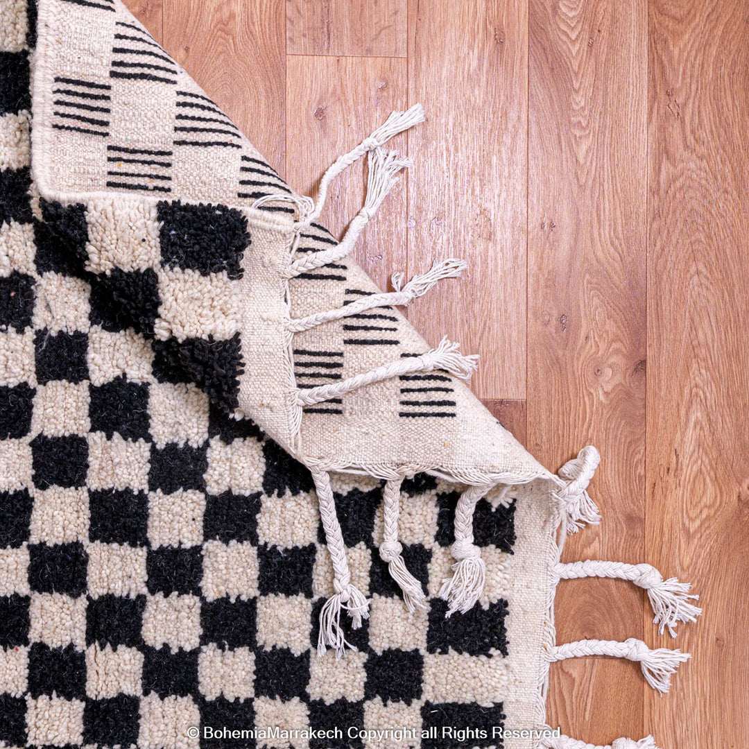 Black and white checkered Moroccan rug with fringes on wooden floor, handcrafted wool, traditional Berber design.