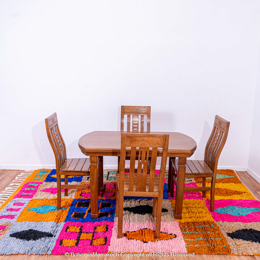 Colorful Moroccan wool rug with tribal motifs under a wooden dining set, featuring pink, blue, orange, and black geometric blocks.