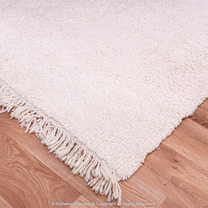 Large white Beni Ourain Moroccan rug on wooden floor, made of natural wool, showcasing fringe detailing.