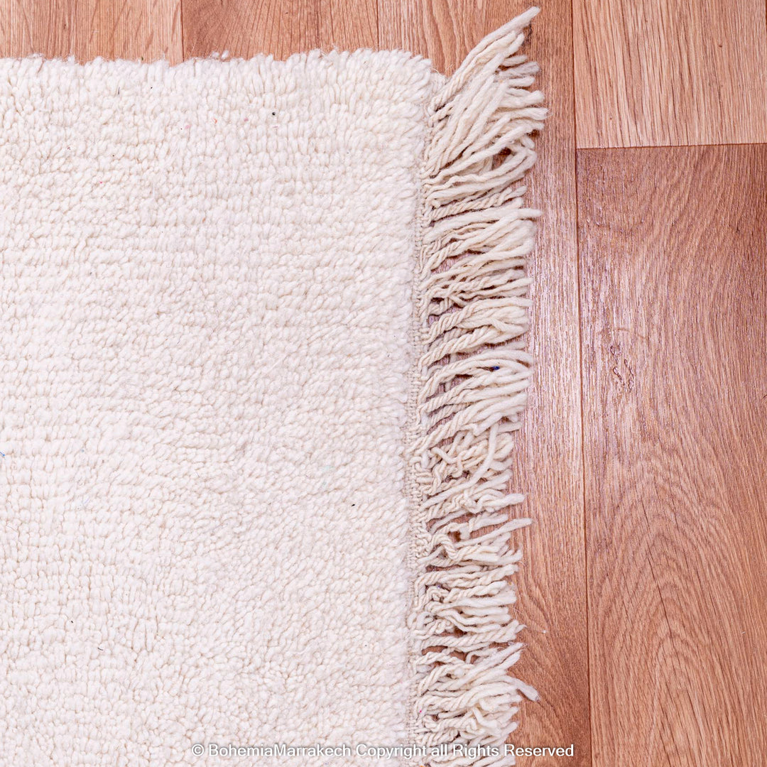 Corner of a plain white Moroccan Beni Ourain wool rug with fringe on wooden floor.