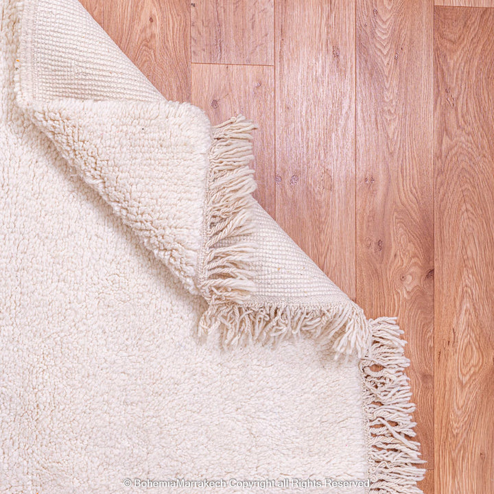 Plain white Moroccan wool Beni Ourain rug on wooden floor, showcasing fringe detail, ideal for minimalist interiors.