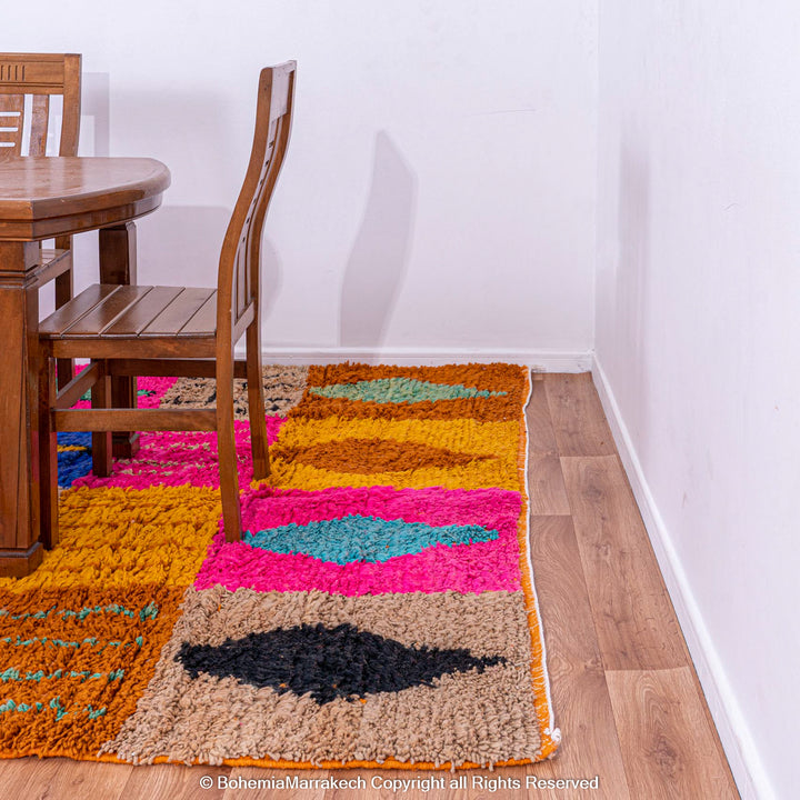 Colorful Moroccan rug with geometric patches under dining table, featuring pink, blue, orange, and black Berber motifs, made of wool.