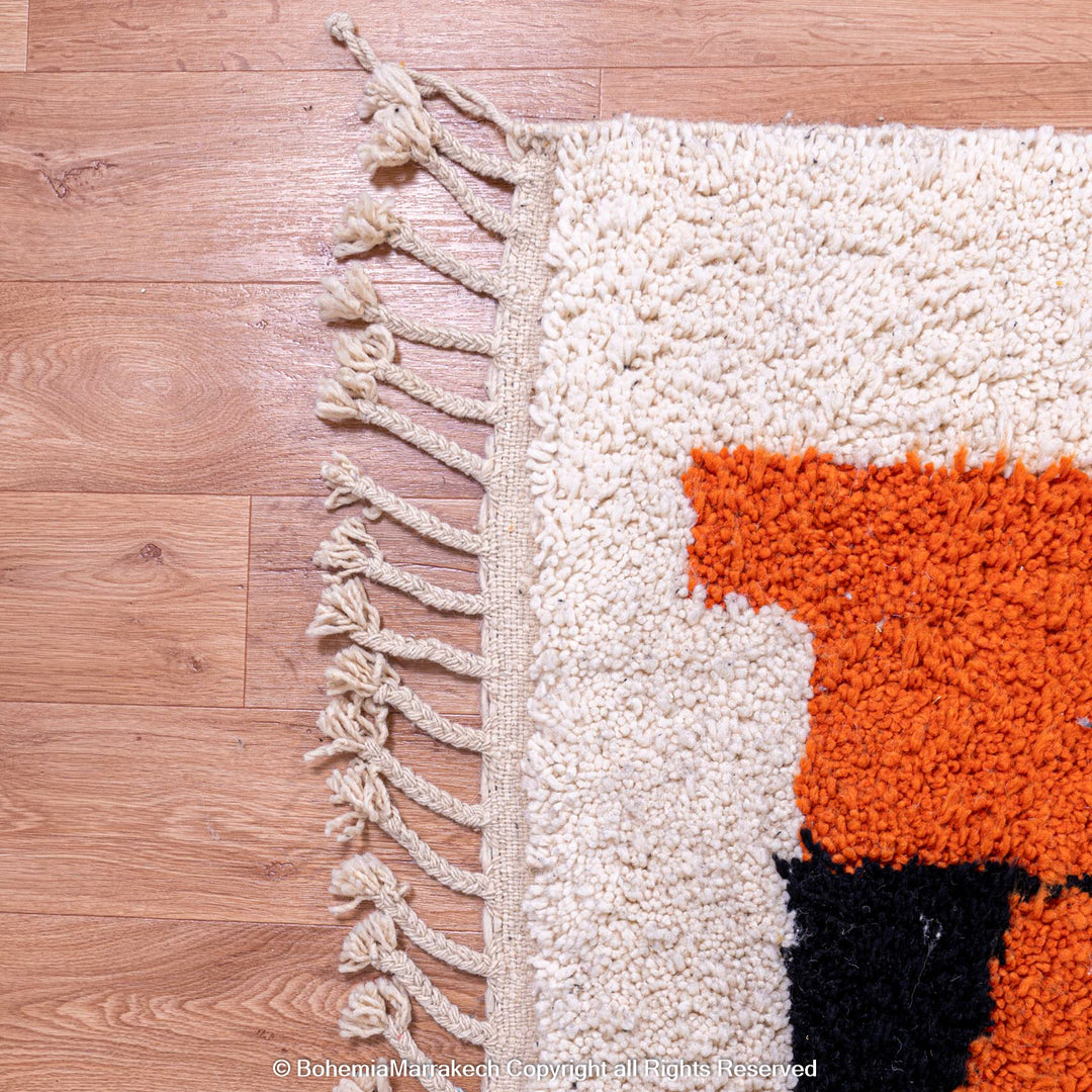 Corner view of handmade Moroccan wool rug with abstract face design in orange, black, and cream tones on wooden floor.