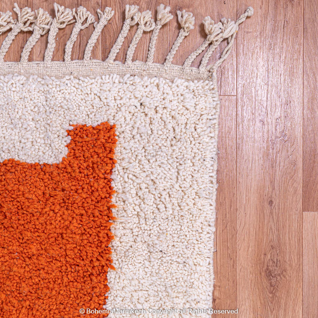 Close-up of handmade Moroccan wool rug with orange and cream design, showcasing Berber craftsmanship and tassel detail on wooden floor.
