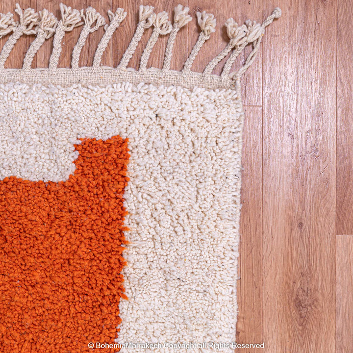 Close-up of handmade Moroccan wool rug with orange and cream design, showcasing Berber craftsmanship and tassel detail on wooden floor.