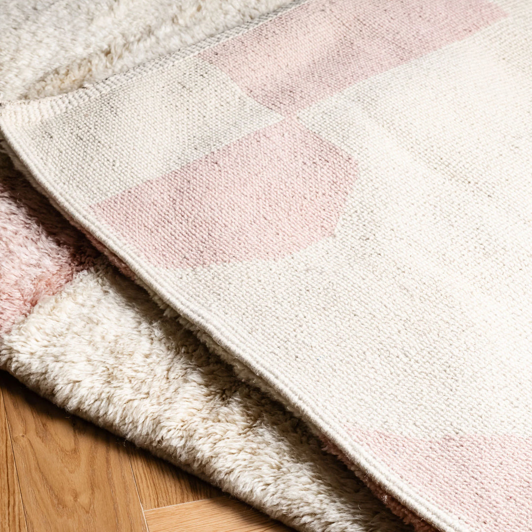Soft pink geometric Moroccan wool rug on hardwood floor, showcasing handmade craftsmanship and cultural design.