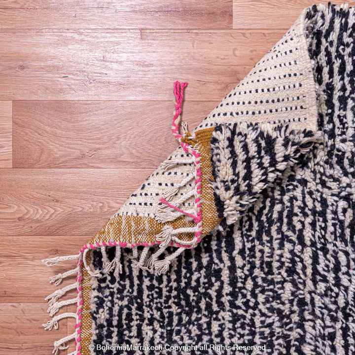 Close-up of a handmade textured Moroccan Berber rug in cream and black with pink, gold, and tassel accents on wooden floor.