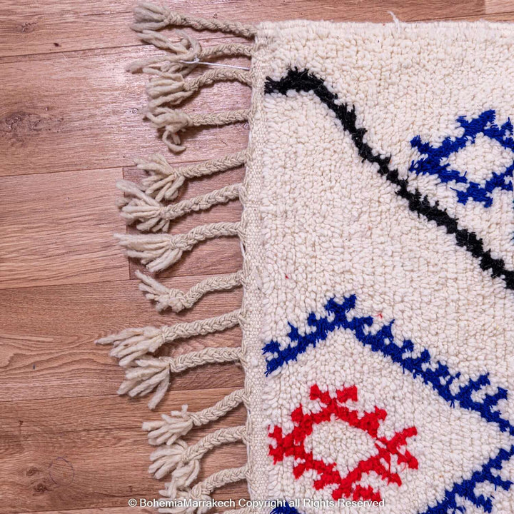 Close-up of handmade Azilal Moroccan rug with red and blue Berber symbols on cream wool, showcasing traditional tribal art.