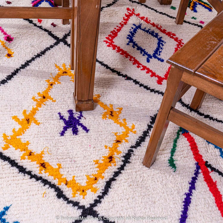 Handmade pink Moroccan Berber rug with colorful embroidered symbols, tribal art, and a cream wool background under wooden furniture.
