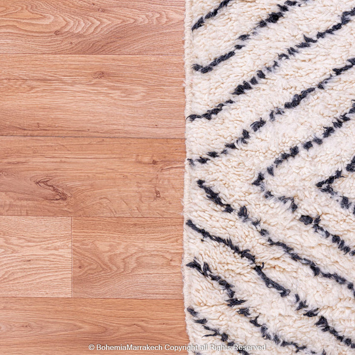 White Moroccan wool rug with black diamond lines on wooden floor, handmade by Berber women in Atlas Mountains.