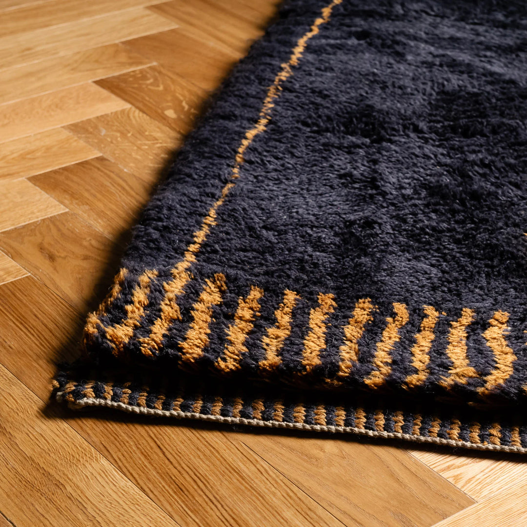 Dark charcoal Moroccan rug with golden pattern on a wooden floor, showcasing handcrafted Berber design and abstract grid.