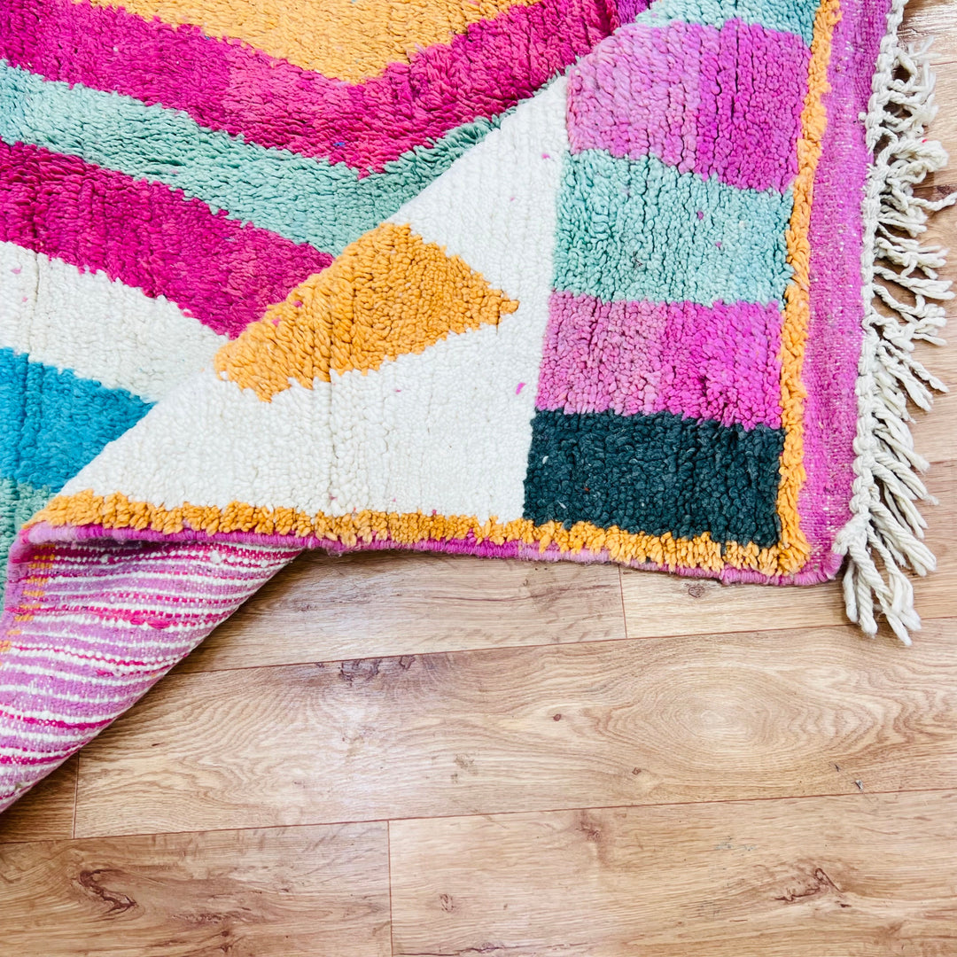 Colorful vintage Moroccan rug with zigzag motif and fringes, featuring orange, teal, fuchsia, and cream tones on a wooden floor.
