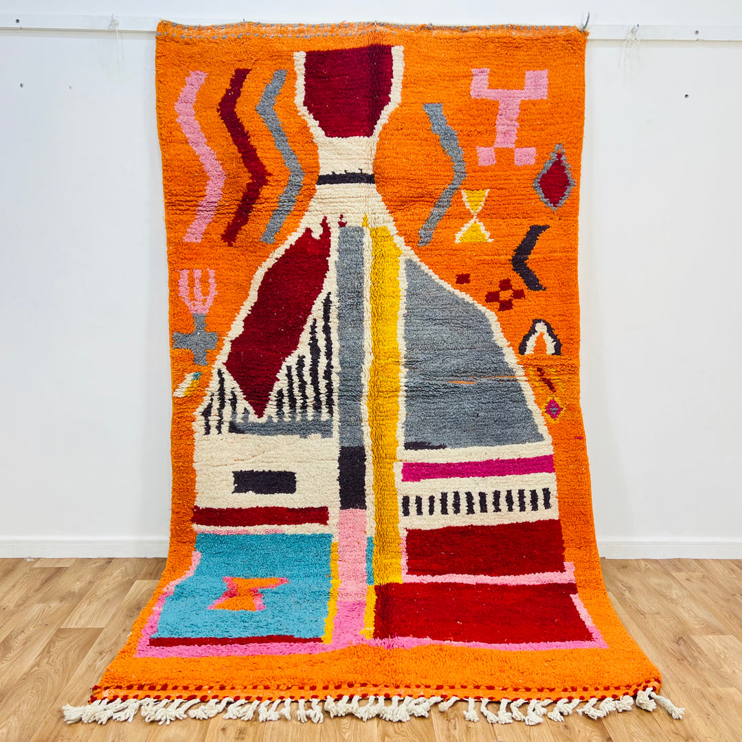 Vintage Moroccan wool rug in vivid orange with abstract tribal patterns, featuring blue, pink, and red tones. 9x5 ft. Handmade Berber rug.