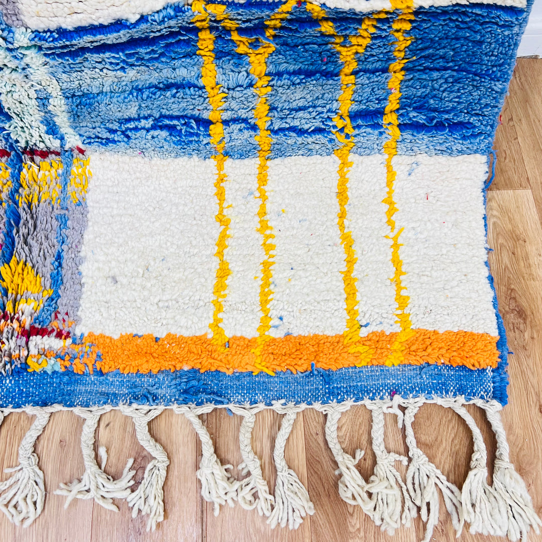Color block Moroccan Berber rug with blue, yellow, and orange patterns on wood flooring. Handwoven wool, 244x168 cm.