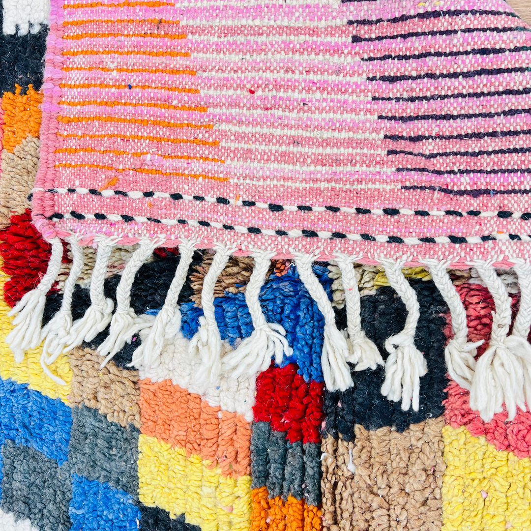 Close-up of a colorful Moroccan rug featuring geometric patterns in magenta, saffron yellow, blue, and pink with fringe detail.