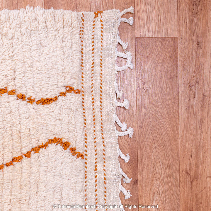 White Moroccan rug with rust-orange diamond pattern on wooden floor, handcrafted wool by Berber artisans, 8x10 area rug.