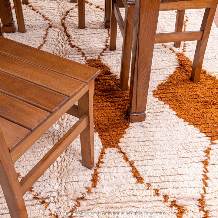 White Moroccan wool rug with rust diamonds under wooden furniture, handmade by Berber artisans, featuring geometric earthy tones.