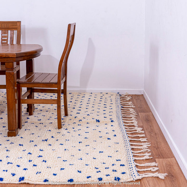 Large white Moroccan rug with blue dots under a wooden dining table, made of handwoven wool in traditional Beni Ourain style.
