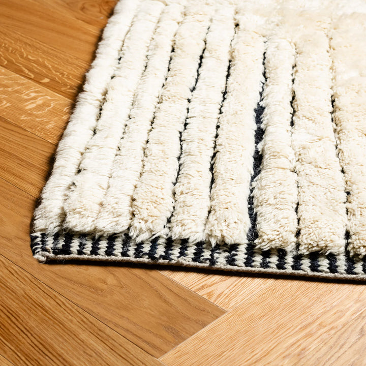 Close-up of a handmade Moroccan Beni Mrirt rug with thick pile and striped geometric design on a wooden floor.