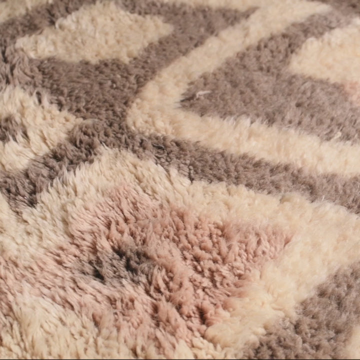 Close-up of a handwoven geometric Moroccan wool rug with diamond patterns in soft pink and beige tones, showcasing texture and detail.