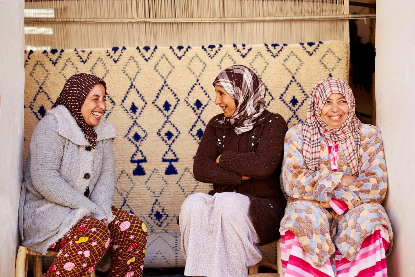 Three Berber women sitting with handmade Moroccan rugs in the background