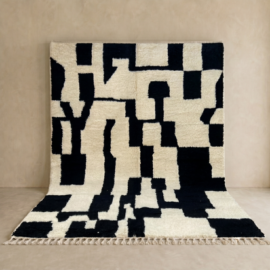 Black and ivory handmade Moroccan wool rug with bold abstract geometric pattern and fringed tassels, displayed against a neutral beige wall.