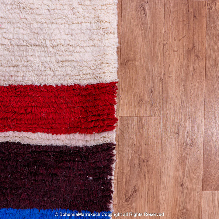 Colorful Moroccan Berber wool rug with red, blue, and ivory stripes on a wooden floor, perfect for eclectic spaces. Moroccan shag rug detail.