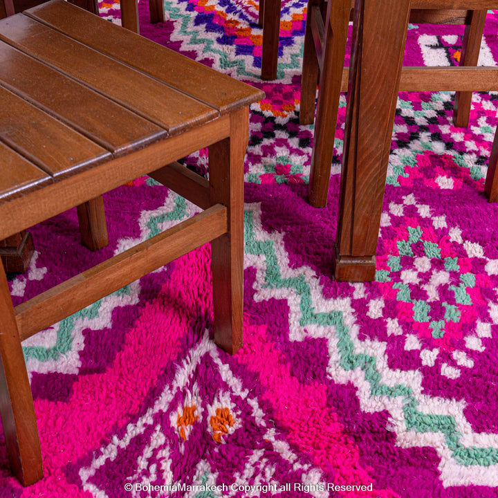 Pink Azilal Moroccan rug with vibrant geometric patterns under wooden chairs, featuring turquoise, violet, and orange motifs.