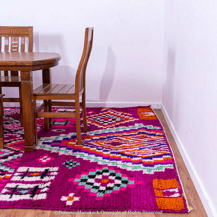 Pink Azilal Moroccan rug with colorful tribal motifs under wooden dining set, showcases vibrant Berber wool craftsmanship.