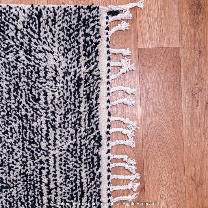 Close-up of handmade grey Moroccan Beni Ourain wool rug on wood floor, showcasing intricate patterns and fringed edges.