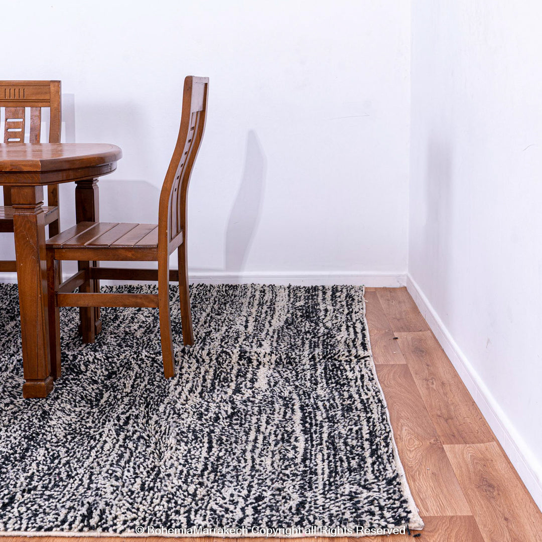 Grey Moroccan Beni Ourain wool rug under wooden dining table with chairs, showcasing marbled dark grey and white tones in room.